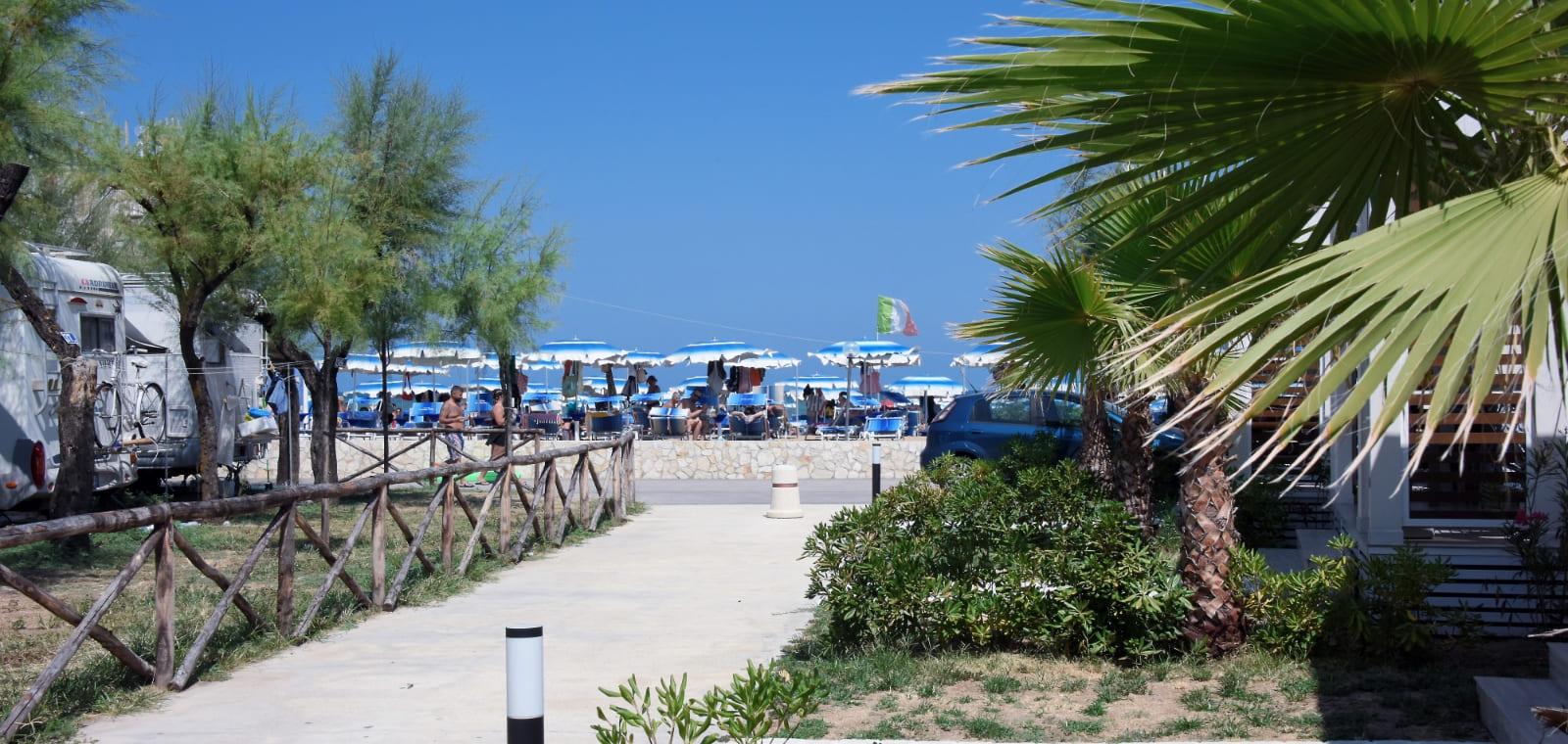 Spiaggia con ombrelloni blu, camper e vegetazione mediterranea, sotto un cielo azzurro.