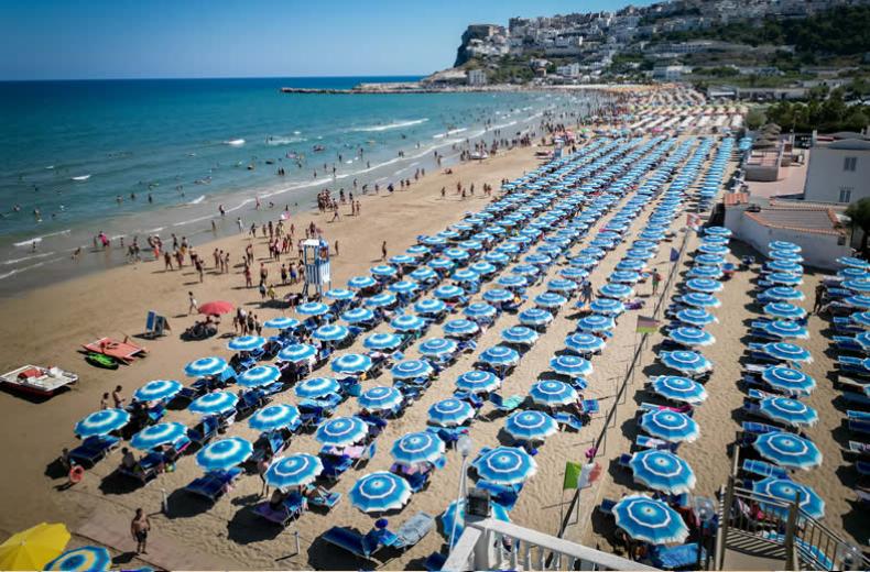 Spiaggia affollata con ombrelloni blu e lettini, mare calmo e cielo sereno.