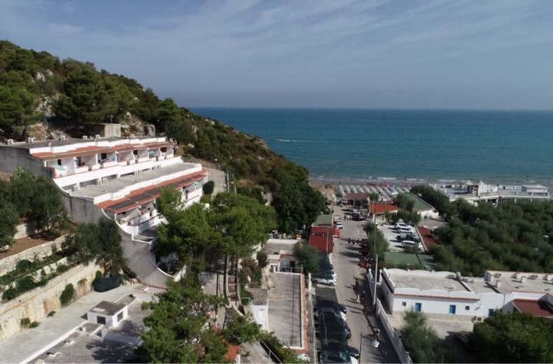 Vista panoramica su un complesso residenziale e la spiaggia, circondati da colline verdi.