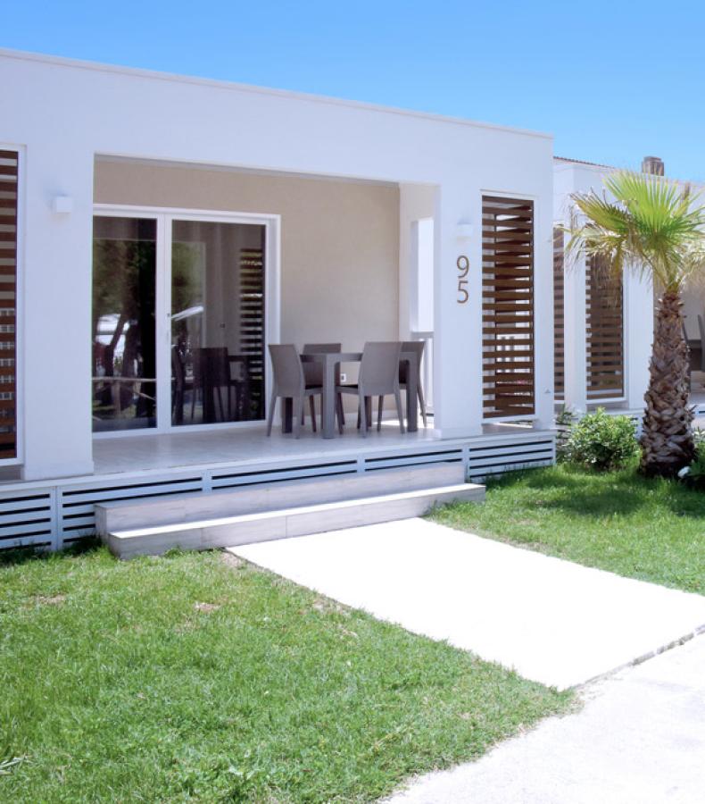 Viale verso la spiaggia con palme e bandiera italiana, ombrelloni blu sullo sfondo.