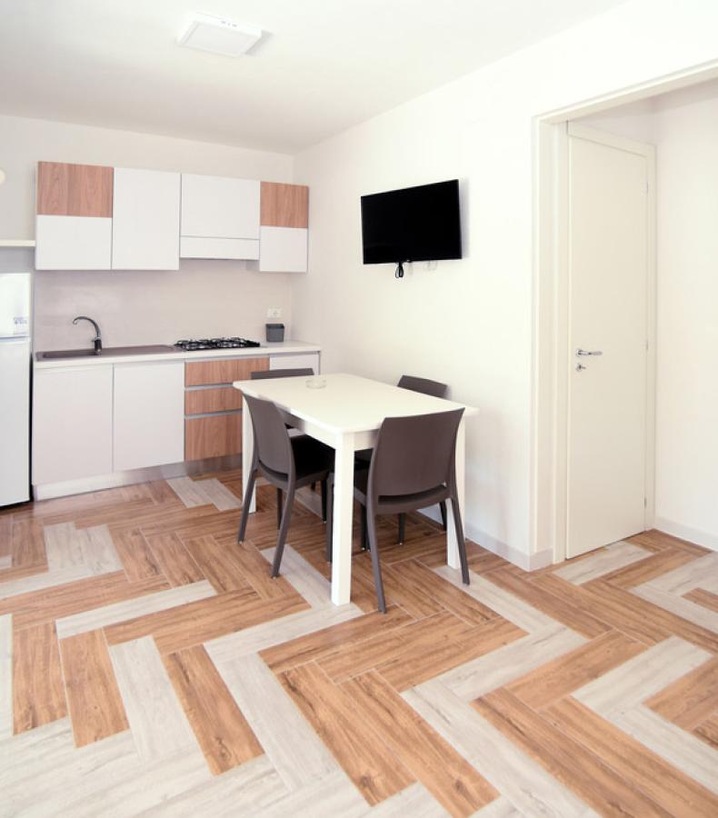 Cucina moderna con parquet, tavolo e sedie, TV a parete, frigorifero e mobili bianchi.