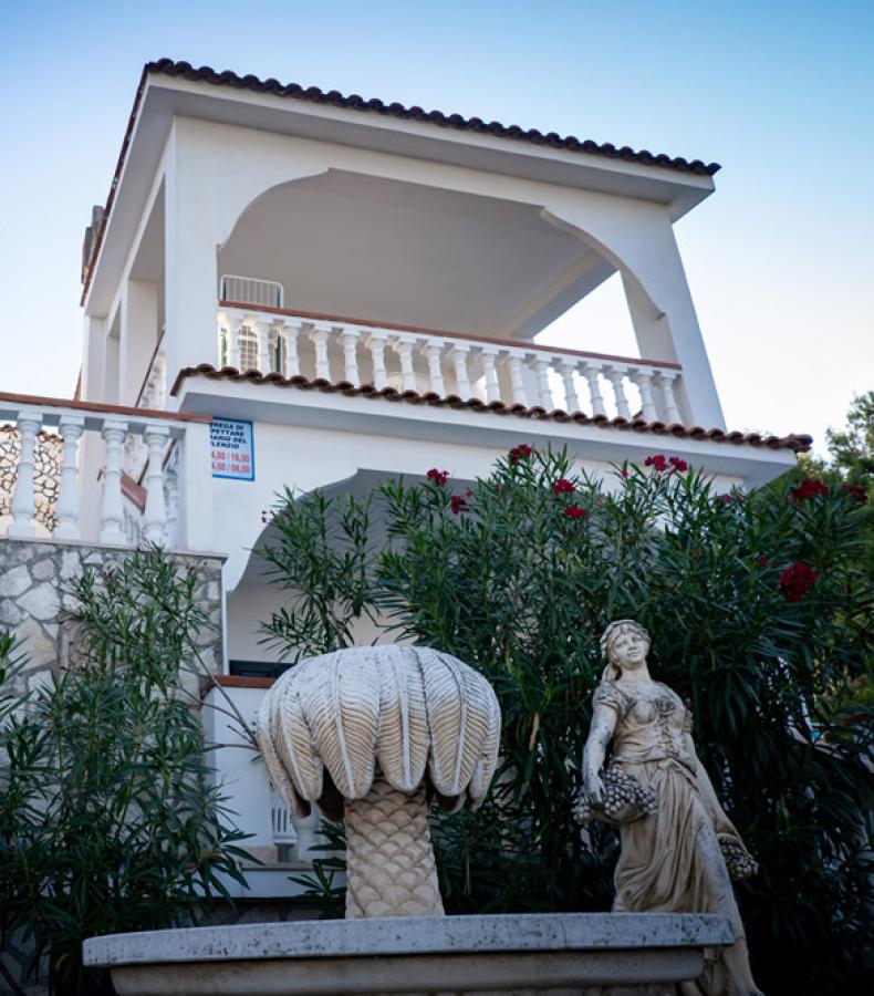 Villa bianca a due piani con terrazze, circondata da alberi e giardino.