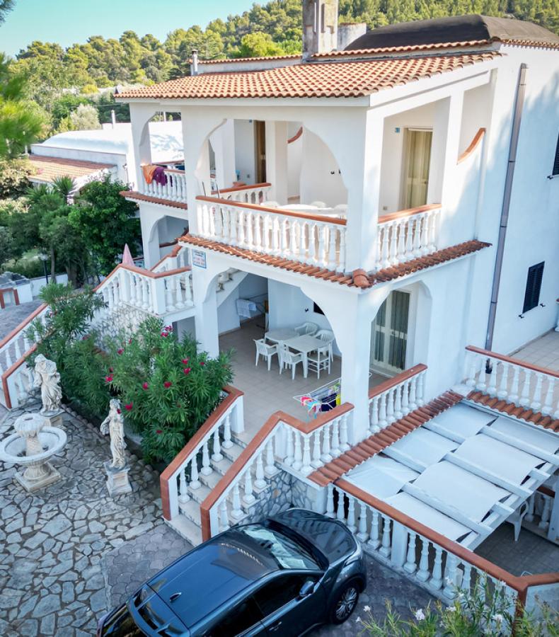 Villa bianca con balconi, giardino, statue e auto parcheggiate.